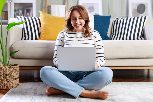 istock Young woman using her laptop while sitting on the floor at home 1870210039