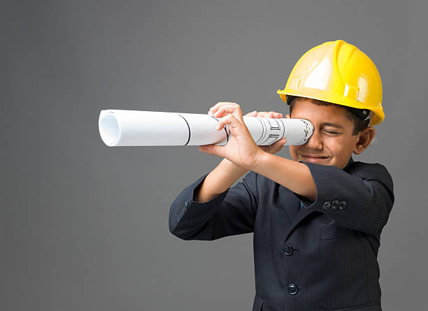 Little boy with yellow hardhat looking through blueprints as handheld telescope