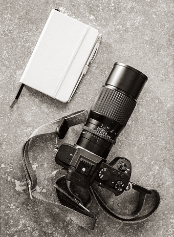 Modern digital camera with telephoto lens and traditional leather-bound paper notebook on granite surface