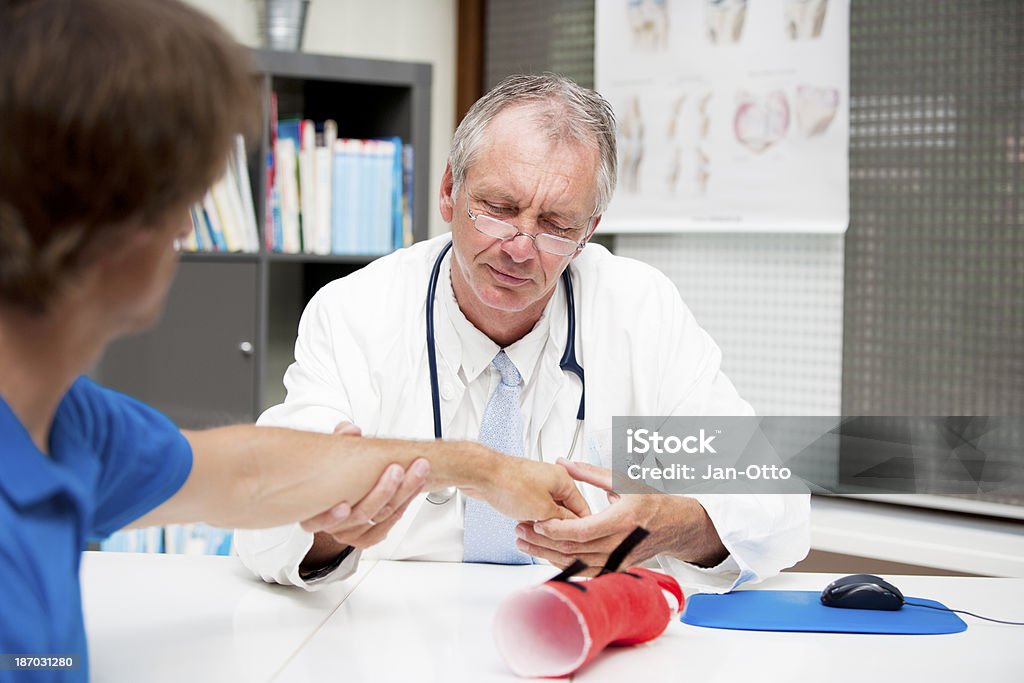 Mature medicine checking lower arm fracture A mature orthopedic medicine, checking the lower arm of a male patient. Maybe the patient had a distal radius fracture. The red cast, spending shelter to the fracture, is lying on the table and had to be taken off during inspection. 50-59 Years Stock Photo