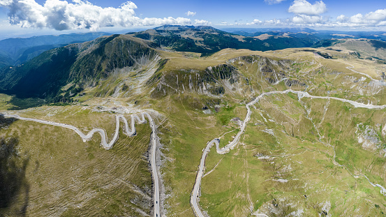 From an aerial view, this image shows stunning mountain roads winding around a calm lake. The dark waters rest among tall mountains, showing off a mix of light and shadows and the roads trail through uninhabited regions. There's also a construction site near a dam.