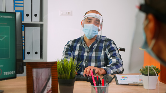 Handicapped businessman with visor and protection mask discussing with colleague working in new normal business office. Man respecting social distance in financial company during global pandemic.