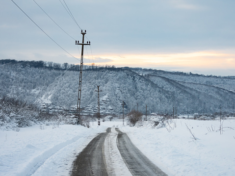 A dirt, country road winding along a snowed field in a hilly rural landscape. Utility poles made from concrete shafts are following the road. Sunset time over the rural area.