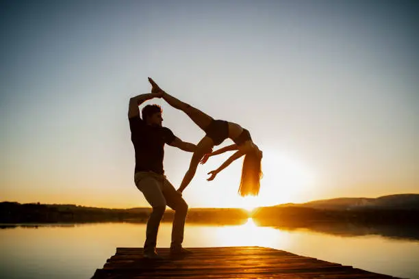 Young man and woman doing acroyoga pose by lake, strength, trust, perfect synchronization, balance and harmony
