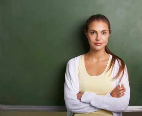 A lovely young teacher standing in front of a blackboard with her arms folded - copyspace