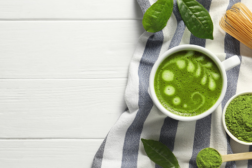 Delicious matcha latte, powder, leaves and whisk on white wooden table, flat lay. Space for text