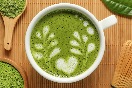 Delicious matcha latte, powder, leaf and whisk on bamboo mat, flat lay