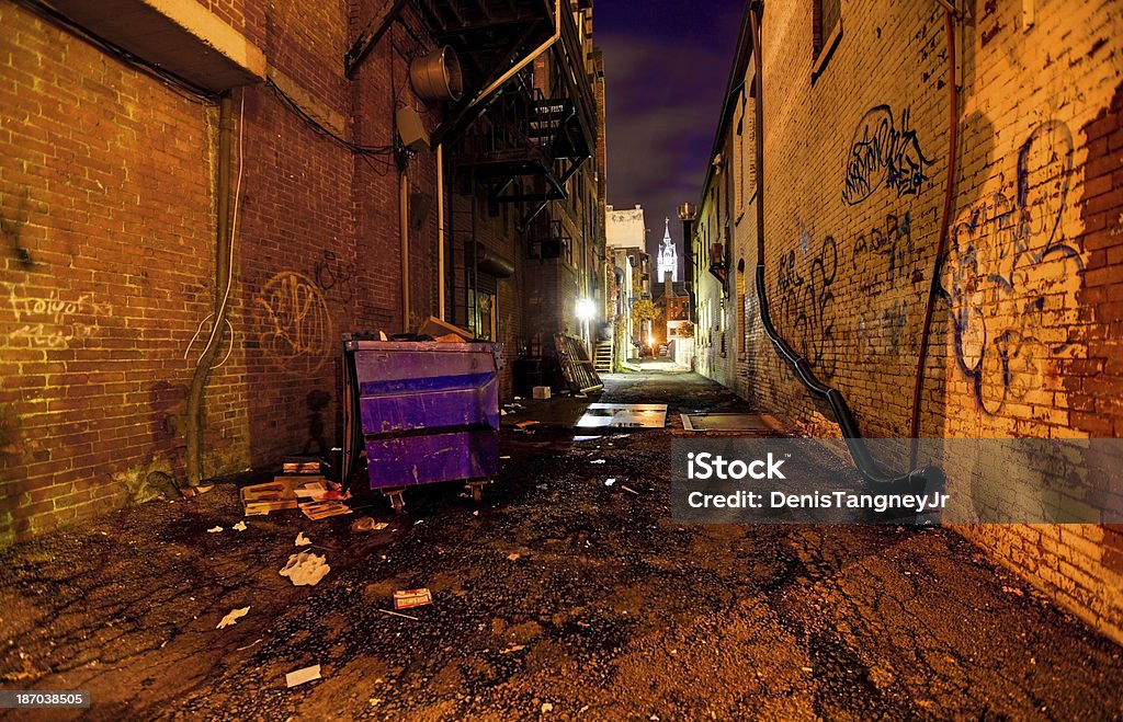 Gritty Dark Urban Alleyway Long dark gritty alley between two old derelict buildings. Trash littered across the pavement with eerie shadows. Uninviting graffiti brickwork walls frame the  dirty and dangerous inner city alleyway Alley Stock Photo