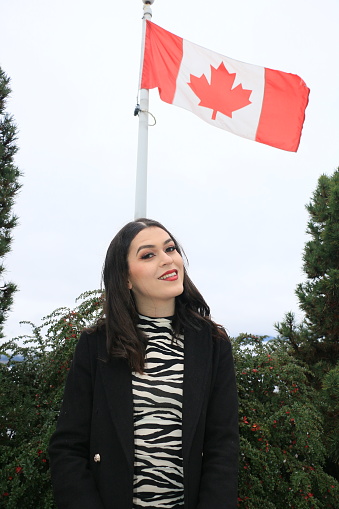 A Mexican immigrant woman in front of a Canadian Flag. She is wearing medium length black hair, makeup, a black unbuttoned jacket and a black and white dress.