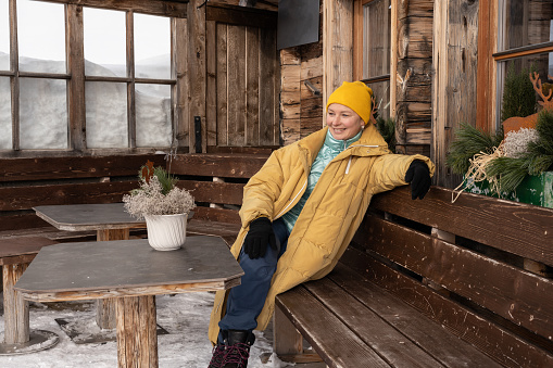 Middle-aged woman in warm clothes sitting with smile on wooden bench near wooden house in winter. High quality photo