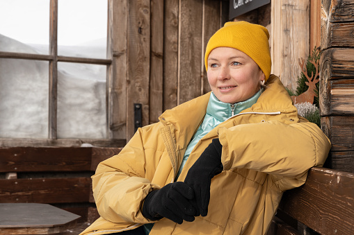 Close-up portrait of a middle-aged woman in winter clothes sitting on a bench near a house against the background of a frame with glass and a snowdrift. High quality photo