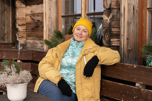 Woman in winter clothes sitting on a bench near a wooden house on a winter day. High quality photo