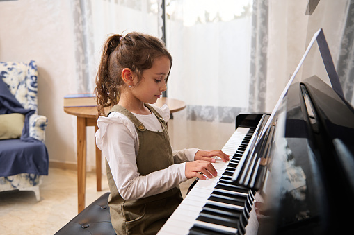 Caucasian little child girl playing piano, sitting alone, touching keyboard and feeling the music rhythm with touching white and black keys, in cozy home interior. Music lesson