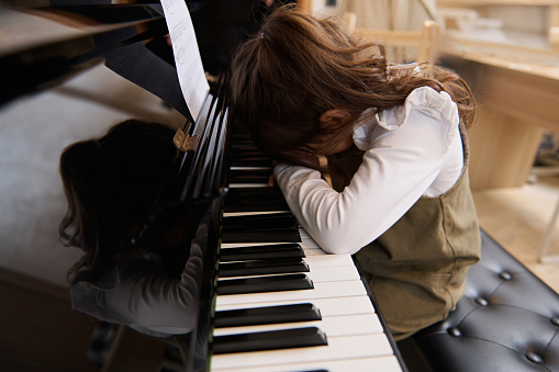 Little kid girl folding her hands and hiding her face, putting her head on piano keys, expressing negative emotions having difficulties or forced to learn playing piano. Childhood. Negative experience