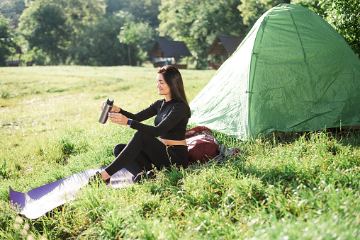 European sport girl sit on fitness mat near tent and drinking tea. Green meadow. Young pretty woman wear sportswear and enjoying sunlight. Concept of resting and tourism on nature. Camping vacation