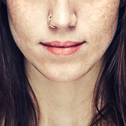 A beautiful close up portrait of an unrecognizable young woman's face from the nose to lower neck.  Natural makeup and a nose ring.  She has brown hair and freckles.  Square crop.