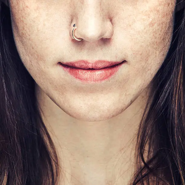 A beautiful close up portrait of an unrecognizable young woman's face from the nose to lower neck.  Natural makeup and a nose ring.  She has brown hair and freckles.  Square crop.