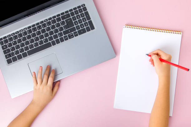 istock Top view of female hands writing in notebook while using laptop on pink background 1870491043