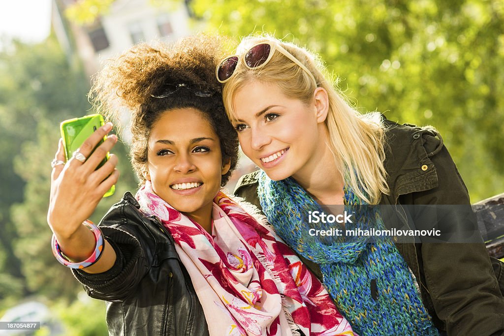 Two girls taking pictures with a Smartphone Two young girls in a park taking pictures with a Smartphone on a sunny day and making funny faces. Adolescence Stock Photo