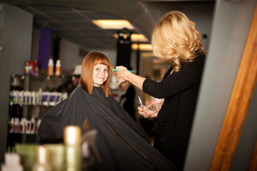 Color image of a little red-haired girl getting her hair cut by a young woman in a salon.