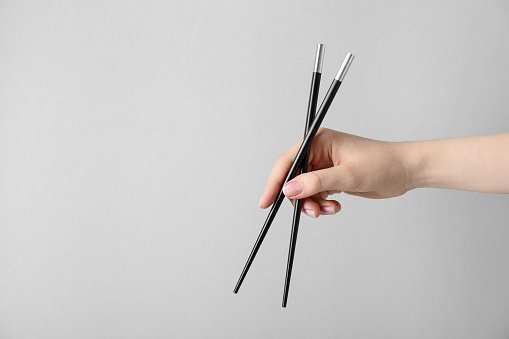 Woman holding pair of black chopsticks on light grey background, closeup. Space for text
