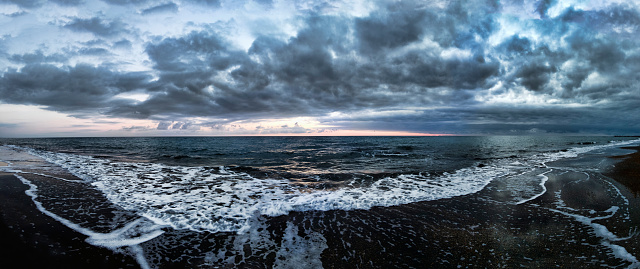 Stunning panoramic 180 degrees of rough sea at sunset on blue hour at beach, a dramatic sky with amazing light and cloudscape and sun reflections with on water edge crashing with foam.
