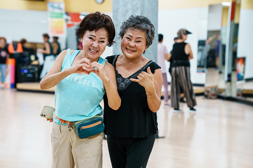 Portrait of two active and healthy senior women of Korean descent smiling directly at the camera after attending a group fitness class together in Hawaii. One woman is making a heart shape with her hands while the other woman is throwing the shaka sign.