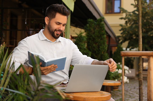 Handsome young man with book working on laptop at table in outdoor cafe