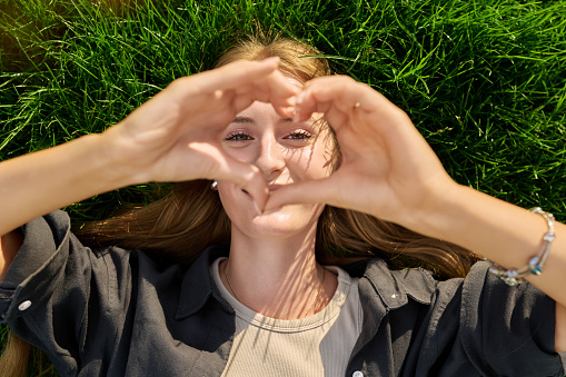 Young happy loving woman showing heart sign with fingers, looking at camera while lying on grass, top view. Happiness love joy emotions