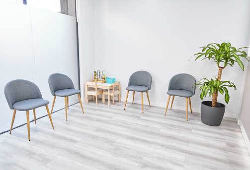 Interior of light waiting room with comfortable gray chairs and small wooden table with various toys for kids near green plant