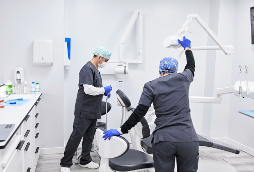 Dentist and assistant in uniform and gloves standing near dental chair and preparing instruments for treatment in modern clinic