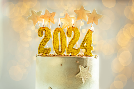 Big golden New Year cake decorated with golden stars and candles against bokeh lights background. Happy New Year 2024. Winter holiday background