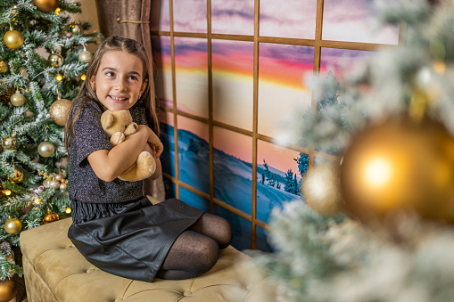 Cute girl hugging teddy bear and sitting on the sofa near the window and Christmas trees.