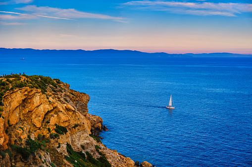 Lonely sailboat or small yacht navigating in harbour of Aegean sea near cape Sounion, place of ancient ruins of temple of Poseidon, on sunset. Beautiful seascape and high cliffs, deep blue sea, distant hazy islands, colorful clouds in sunset sky.