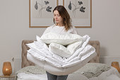Woman holding pile of soft white folded duvet and pillows at home in her bedroom, cozy domestic lifestyle, housewife cleaning, tidying up bedroom, housework chores concept.
