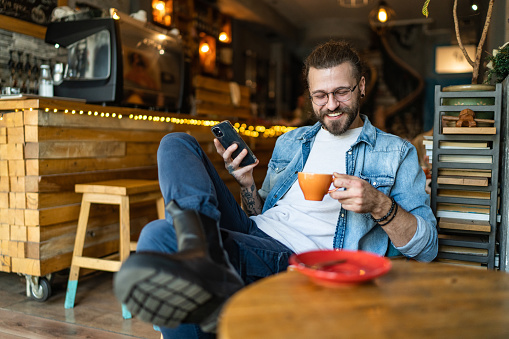 Smiling young man using phone while enjoying in coffee at coffee shop