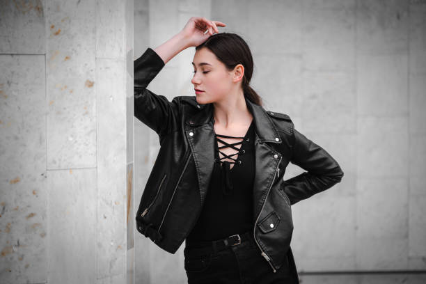 Portrait of a black haired young girl in black clothes and a leather jacket posing thoughtfully against the marble background of the city wall.