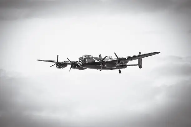 An aged image of an Avro Lancaster bomber in flight.  Black and white photo with vignette for ageing effect.  Made famous for the Dambuster raid on the Ruhr Valley by 617 Squadron of Air Marshal Sir Arthur Harris's RAF Bomber Command, commanded by Wing Commander Guy Gibson 1943.  Lancaster is flying diagonally towards the camera from right to left.  Background is clear with the aeroplane in the center of the image.
