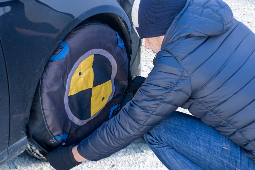 Man installing textile snow chains or socks on the car wheel to avoid skidding. Traction and tire grip for safety driving on winter travels.