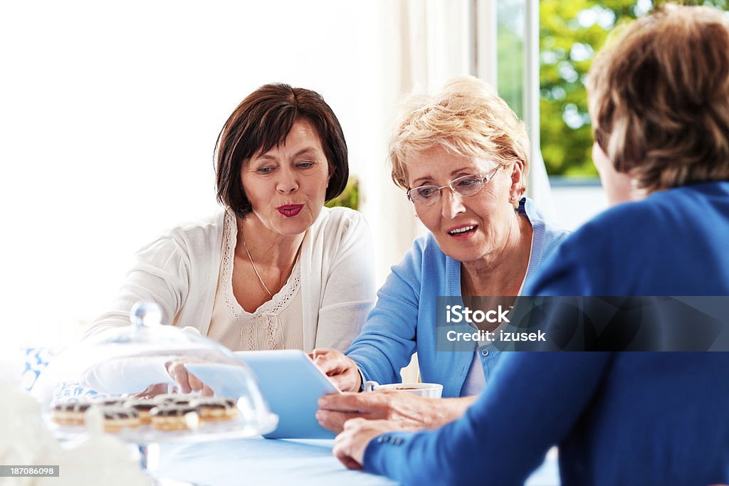 Mature women using digital tablet Three cheerful mature women sitting at the table at home and using a digital tablet together. Lifestyles Stock Photo