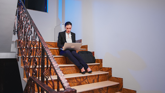 Exhausted overworked entrepreneur doing overtime on project deadline typing on laptop. Serious entrepreneur working on corporate job sitting on staircase of business building late at night.