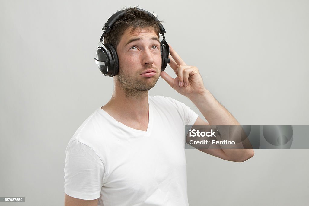 Young Man with Headphones listens to something boring. Young man with casual white T-Shirt and heandphones making disapproving, bored face. Grey studio background. Adult Stock Photo