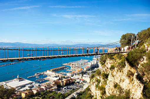 Gibraltar - 11th November 2023: Young adults crossing the Windsor Suspension Bridge (2016) in the Upper Rock Nature Reserve on the Rock of Gibraltar.  Below them is part of the west coast of the city, showing Mid Harbour Marina, Coaling Island Boat Harbour, The Island, Europlaza and Queensway Quay. The town of Algeciras in Spain is in the background, beyond the Bay of Gibraltar.