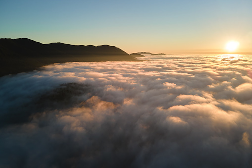 Aerial view of colorful sunrise over white dense fog with distant dark silhouettes of mountain hills on horizon.