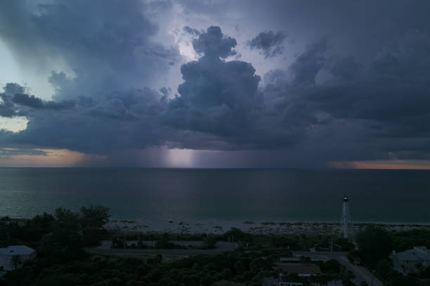 ciudad costera con casas oscuras y faro en la orilla del océano con luz parpadeante en la noche de tormenta para la navegación de embarcaciones comerciales. tormenta eléctrica con relámpagos sobre el agua de mar que representan un peligro para los barc - small town america flash fotografías e imágenes de stock