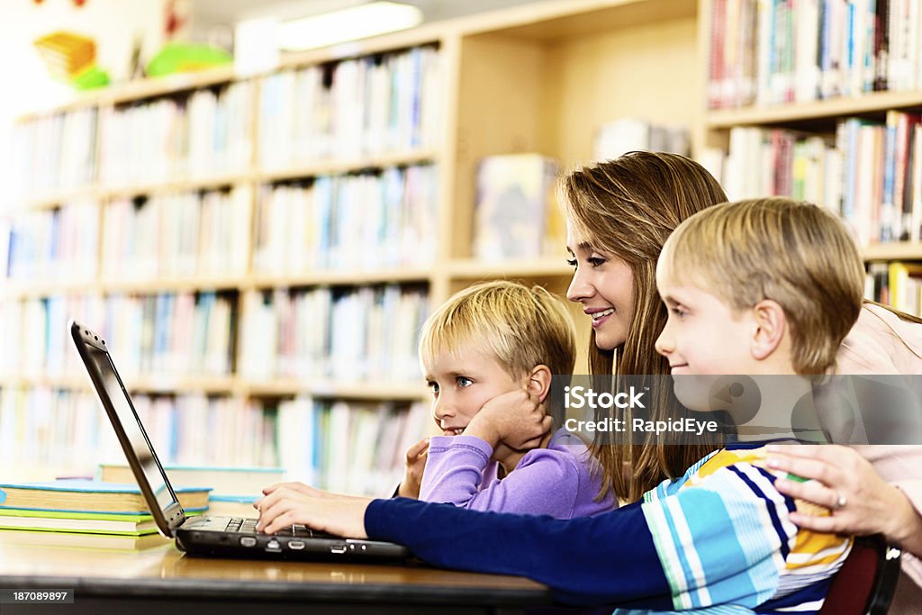 Mulher sorridente ajuda a crianças com o laptop na biblioteca - Foto de stock de 10-11 Anos royalty-free