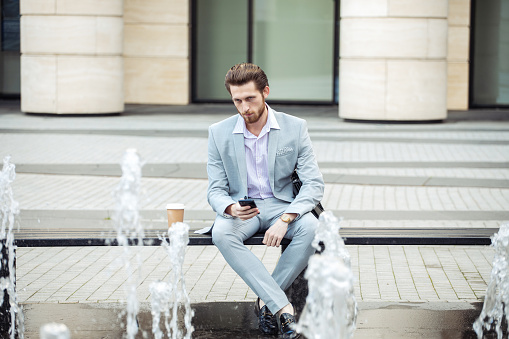 A young businessman sits by a fountain with coffee and a smartphone. Lifestyle and business concept.