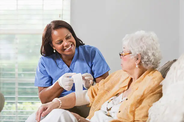 Senior Hispanic woman (70s) with Hispanic healthcare worker (50s) wrapping arm in bandage.  Focus on healthcare worker.