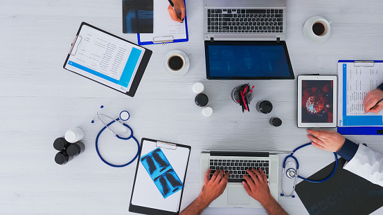 Top view of doctors discussing about virus using tablet, taking notes sitting on desk in medical clinic with digital technology. Assistants writing on laptop and clipboard, researching on copy space