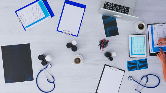 Top view of medical practitioner checking appointments using tablet, tacking notes on clipboard sitting on clinic desk with x-ray and medical equipment all around. Copy space, flat lay concept.